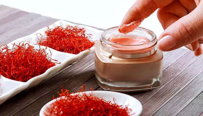  زعفران را در یک ظرف شیشه ای زیبا مشاهده می کنید. ظرف شیشه ای شفاف است و رنگ زعفران را به زیبایی به نمایش می گذارد. زنی در حال استفاده از کرم زعفران برای داشتن پوستی درخشان است
